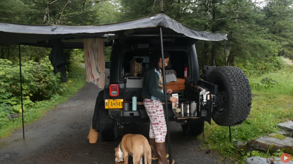 A woman stands at the back end of her Jeep Wrangler gathering supplies while her dog sniffs around